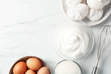 Delicious homemade meringue cookies, ingredients and whisk on white marble table, flat lay. Space...