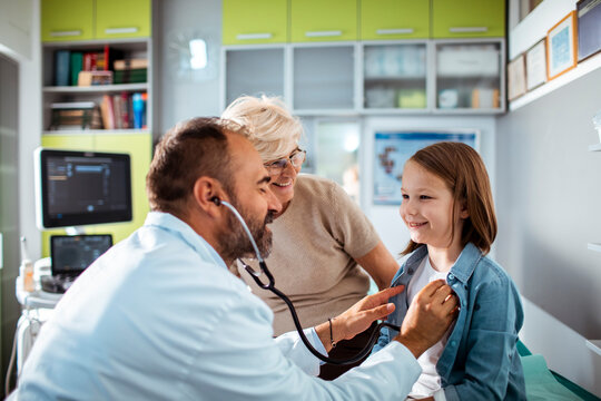 Doctor examining young girl with stethoscope while grandmother watches in medical clinic