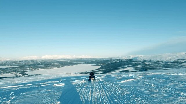Powerful snowmobile gliding down snow covered mountain slope, revealing expansive frozen lake and majestic mountain range under sunny winter sky in pristine Lapland wilderness