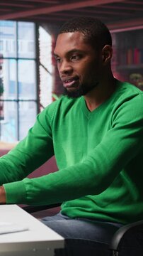 Vertical Video Black man doing remote work feeling uneasy and anxious at desk, shaking his legs and being jittery due to task completion and deadline. Freelancer working under pressure, anxiety