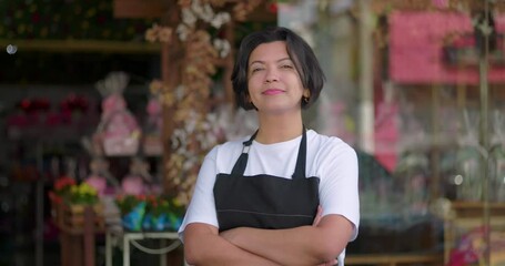 Florist standing with thoughtful expression outside decorated flower shop, arms crossed, showing pride and calm in blooming urban setting full of arrangements and charm