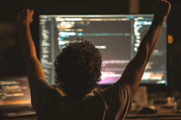 Successful Latino developer celebrates coding success with arms raised in front of program code on computer
