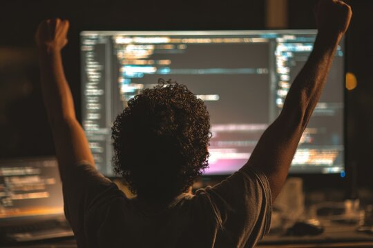 Successful Latino developer celebrates coding success with arms raised in front of program code on computer