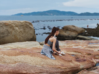 A thoughtful young Asian woman sitting on a rocky beach, dressed in athletic wear, reflecting on life against a serene seascape