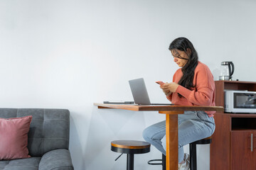Latina woman working from home with smartphone and laptop in kitchen area