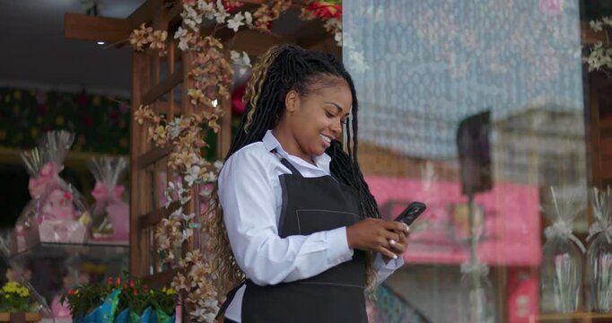 Florist smiling and looking at phone outside flower shop, enjoying break while checking messages near floral arrangements and decorative boutique entrance