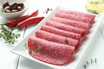 Slices of tasty smoked sausage, spices and olives on white wooden table, closeup