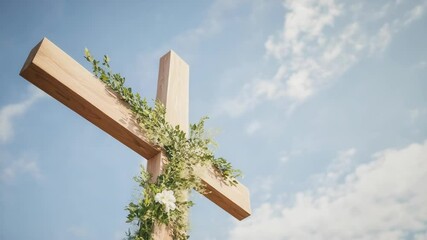 Wooden Christian cross decorated with foliage and white flower against soft blue sky with clouds on a bright sunny day, religion and faith concept.
