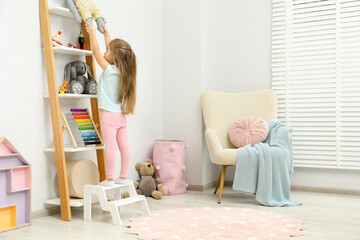 Little girl standing on step stool and reaching for toys on shelf indoors. Space for text