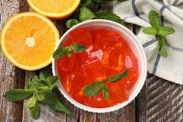 Delicious jelly cubes in bowl, mint and orange on wooden table, flat lay