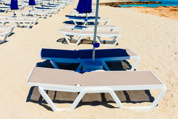 Rows of white beach chairs are neatly arranged on a sandy beach under blue umbrellas, casting shadows in sunlight. In background turquoise sea meets rocky cliffs, creating  serene coastal scene