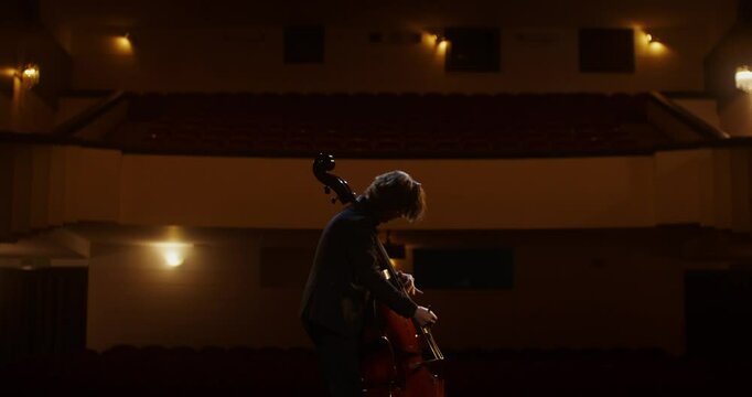 A man plays the double bass using a bow. He stands on the stage of the theater