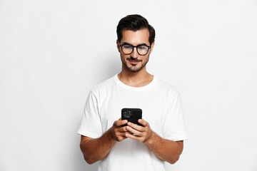 Smiling young man in a white t-shirt using mobile phone, standing against a plain solid white background. Tech lifestyle concept.