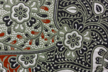 View of a traditional batik fabric showcasing intricate floral and paisley patterns in muted green, black, white, and touches of red. The textile exhibits fine detailing and a rich cultural aesthetic.