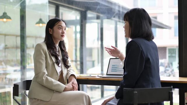 Two women are sitting at a table, one of them is talking to the other. The woman on the left is wearing a suit and the woman on the right is wearing a black suit. They are both looking at a laptop