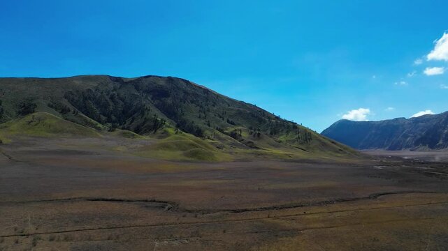 Scenic drone footage panning right to left over the lush green Teletubbies Hill in Bromo. Rolling hills meet a bright blue sky, creating a vibrant and peaceful natural landscape.