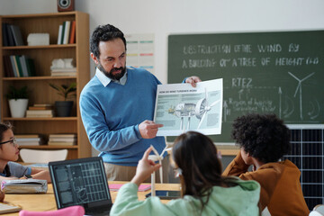 Man explaining wind turbine mechanics to students in classroom with chalkboard in background featuring wind turbine diagrams, engaged in learning environment