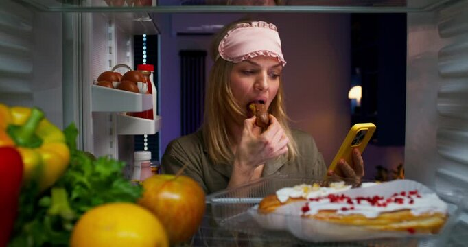 A woman with a sleep bandage on her head eats a cake and uses a mobile phone while standing near an open refrigerator at night. View from the refrigerator