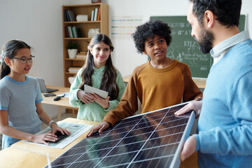 Teacher demonstrating solar panel to attentive diverse students in classroom. Engaged, curious...