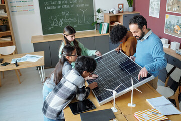 Students gathered around table in classroom examining solar panel and discussing its functionality,...
