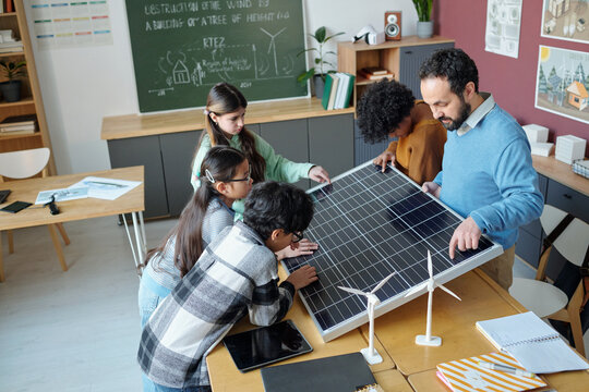 Students gathered around table in classroom examining solar panel and discussing its functionality, guided by male teacher. Chalkboard with diagrams in background