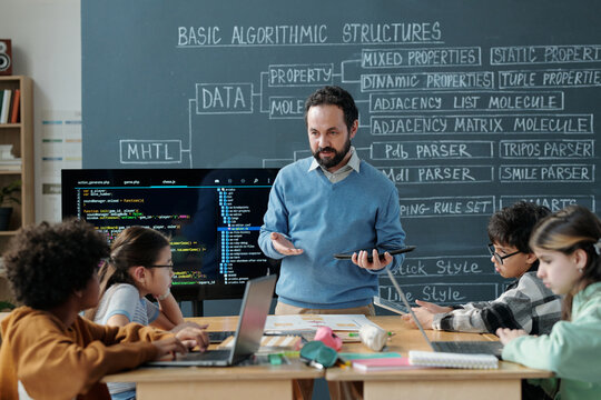 Teacher explaining coding concepts to group of elementary school students seated around table with laptops and textbooks, classroom featuring detailed algorithmic structures on chalkboard