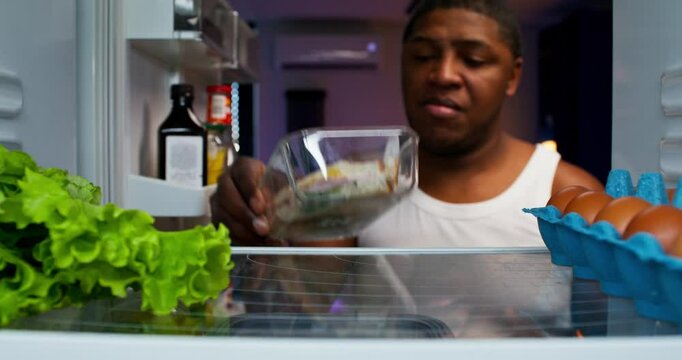 A man eats at night. He opens the fridge and takes a bite of a sandwich. View from the refrigerator