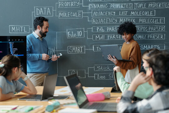 Multiracial group of students learning computer science from a teacher in classroom setting with chalkboard filled with diagrams and terms - Powered by Adobe