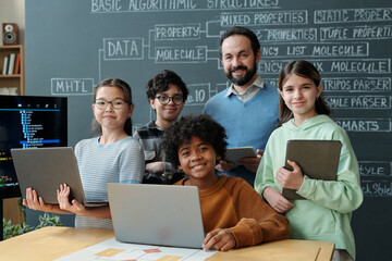 Diverse group of students and their teacher participating in technology class, holding laptops and tablets, and smiling for camera. Visible coding activity on the screen in background