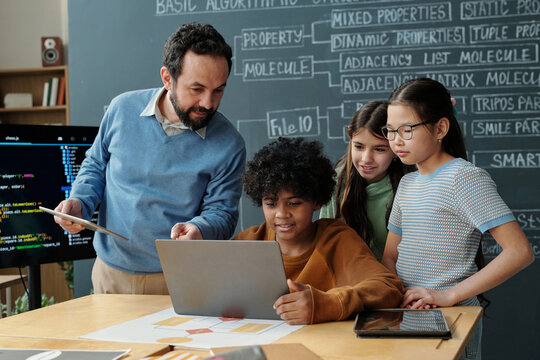 Teacher guiding students in classroom with laptop and tablet, focusing on coding. Students are enthusiastic and collaborative while learning programming and technology