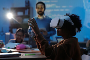 Child engaged in virtual reality learning experience in classroom, with teacher and fellow students...