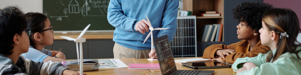 Teacher explaining wind turbine model to diverse group of attentive students in classroom setting, enhancing understanding of renewable energy concepts