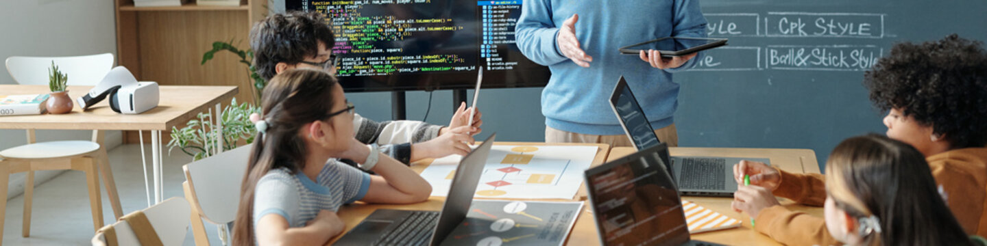 Young students attentively listening to instructor explaining coding concepts using a tablet, surrounded by laptops, books, and digital displays with code in background