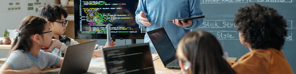 Classroom setting with students attentively listening to teacher explaining coding concepts. Laptops and a screen displaying code are prominent in the learning environment