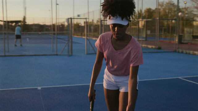 Golden hour illuminates a young black woman tennis player on a blue court as she prepares to serve, other players are visible in the background