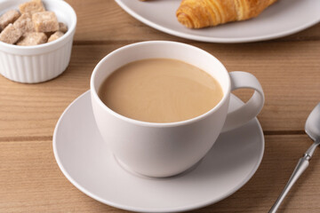 Warm coffee in a white cup served with a croissant and sugar cubes on a wooden table