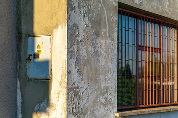 fragment of the old building facade with visible traces of peeling paint. On the left side there is a rusty electrical box, while on the right - a window secured with bars. The sunlight casts a clear 