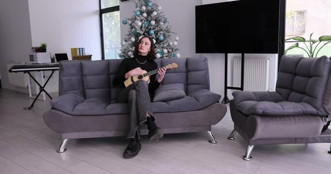 Woman sits cross-legged on grey futon playing ukulele. Female person relaxes in living room during Christmas season given decorated tree slow motion