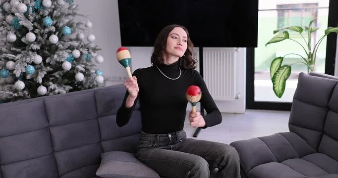 Woman sits on futon holding and shaking maracas with playful expression. Decorated Christmas tree suggests festive and musical atmosphere at home