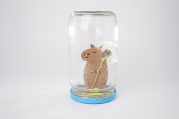 Creative eco-friendly capybara figure holding dandelion inside inverted glass jar with blue lid,...