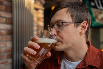 Young man wearing glasses enjoys a refreshing glass of beer at a pub, savoring the moment and the cold beverage in a relaxed and casual atmosphere