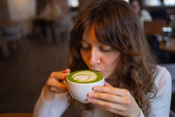 Young woman with long brown hair savors a cup of matcha latte in a cozy cafe, gently blowing on the...