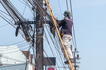 Utility worker repairs power lines using a bamboo ladder in a vibrant urban setting in Thailand