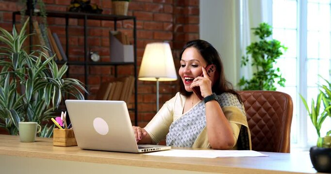 Speaking on smartphone while working at office, Indian young businesswoman in ethnic saree multitasks at desk with laptop, confidently handling work communication using speakerphone in modern setup