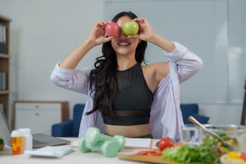 Asian nutritionist smiling while holding two apples over her eyes, sitting at a desk with a laptop, notebook, dumbbells, fresh vegetables, and supplements