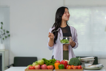 Young woman in fitness attire enjoying a fresh salad in a vibrant kitchen filled with fruits and vegetables, reflecting a healthy and active lifestyle
