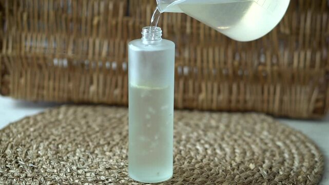 Woman pouring homemade face toner into a transparent frosted glass bottle on beige background. Natural skincare, DIY cosmetics, clean beauty, and minimalist self care concept.