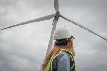 A confident female engineer in a hard hat and safety vest stands proudly before a wind turbine,...