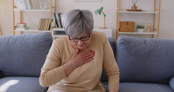 Elderly woman sitting on sofa, clutching chest with both hands, suffering from pain and shortness of breath. Senior lady experiencing symptoms of possible heart attack or infarct and feeling unwell.