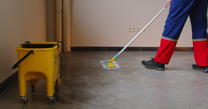 Cropped view of person in work uniform cleaning wet floor with mop in empty room with yellow mop bucket on wheels nearby. Professional janitor performing routine service and floor sanitation work.